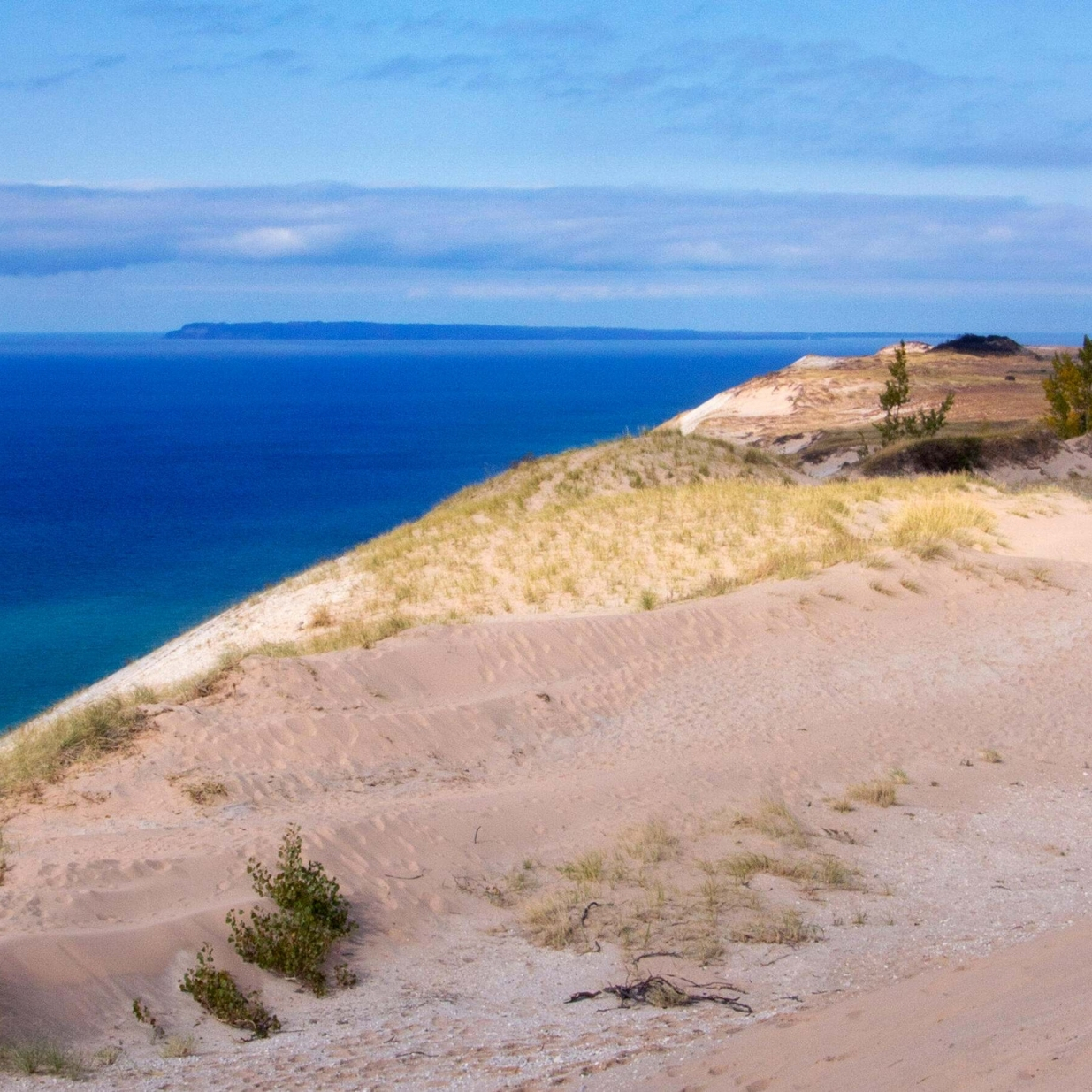 Sweeping sand dunes covered with beach grass overlook a deep blue lake, with sandy bluffs, a distant shoreline, and a partly cloudy sky stretching across the horizon.