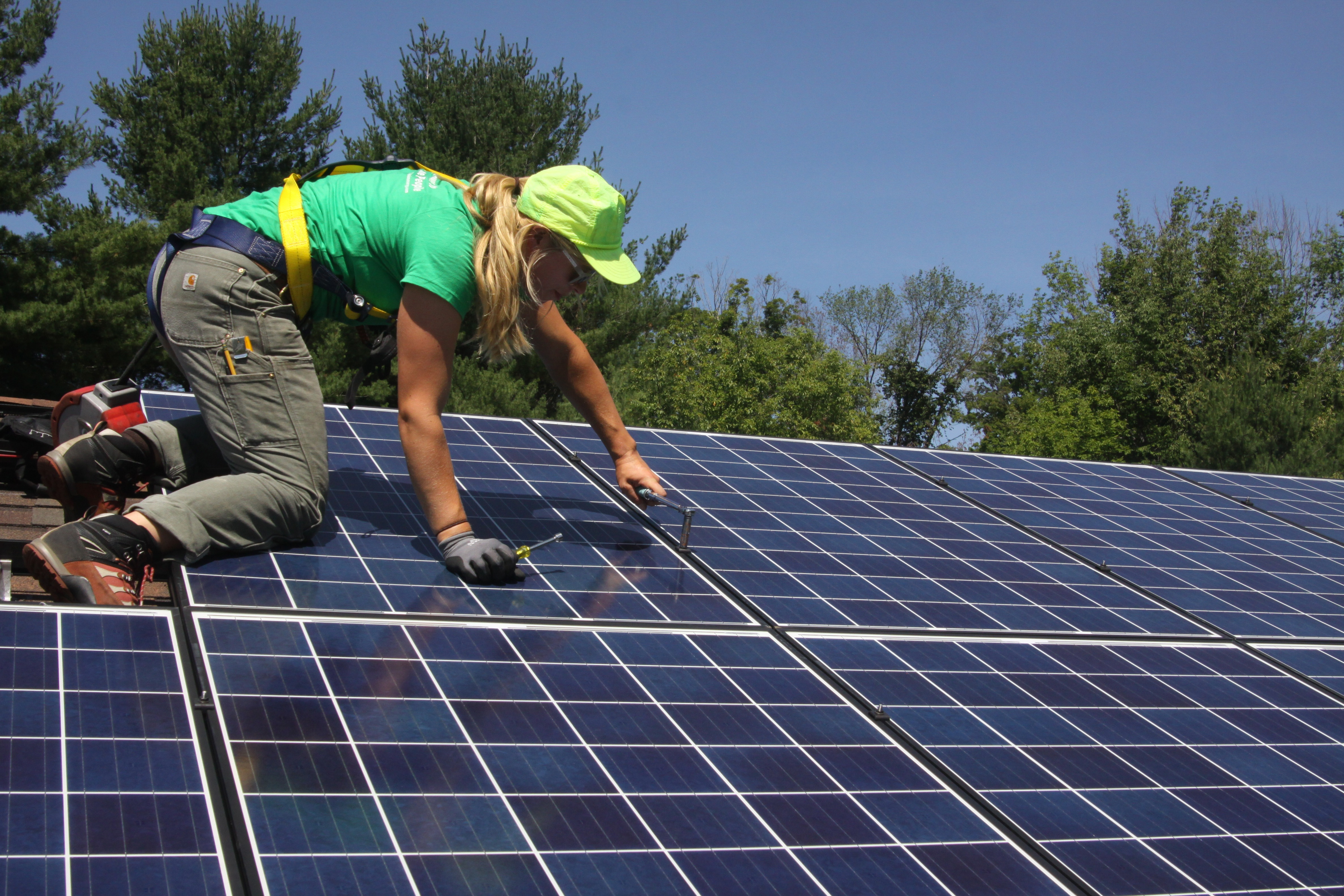 A woman kneels to install solar panels on a roof.