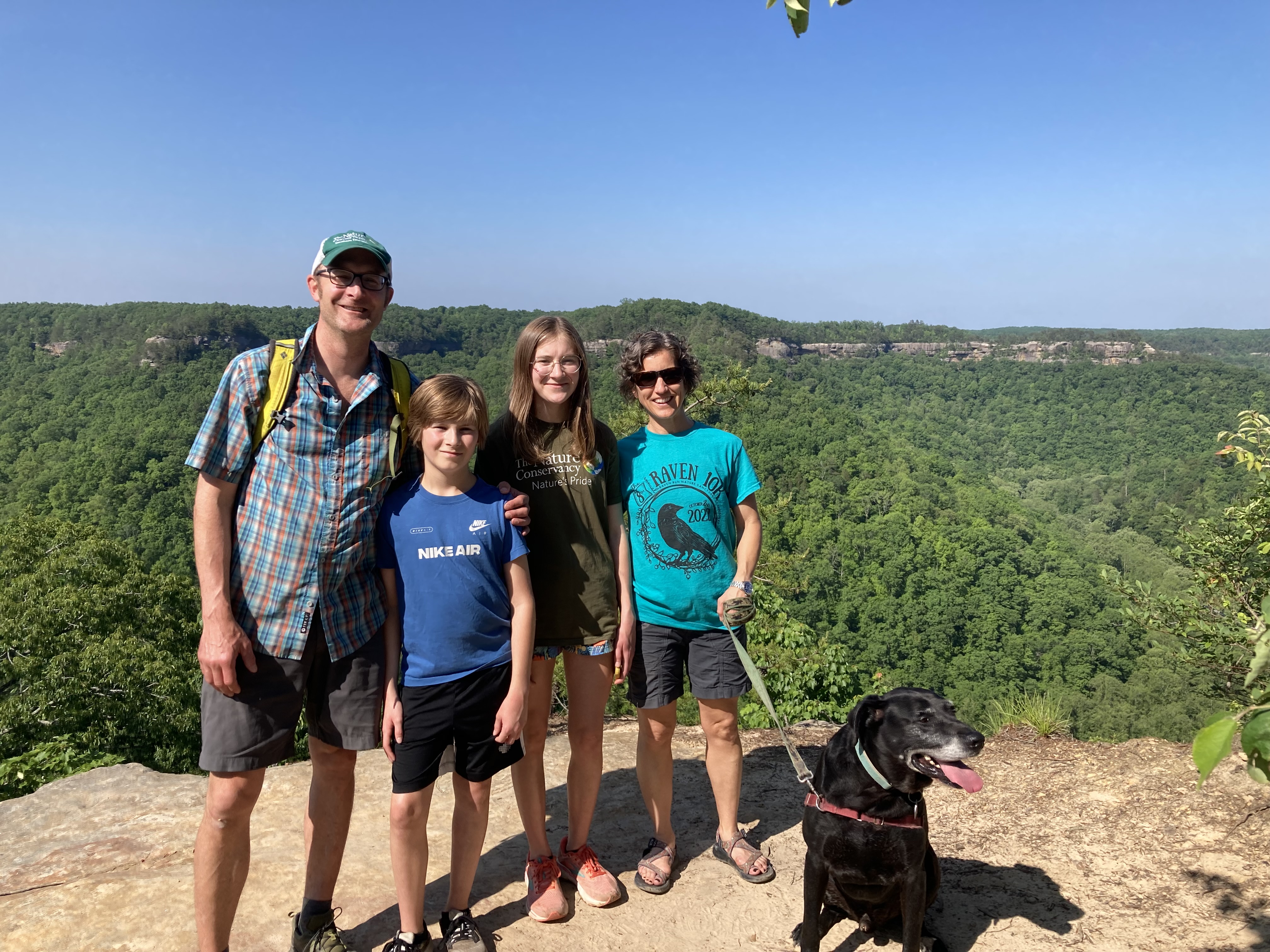 Four family members and a dog smiling together with a green forested view behind them.