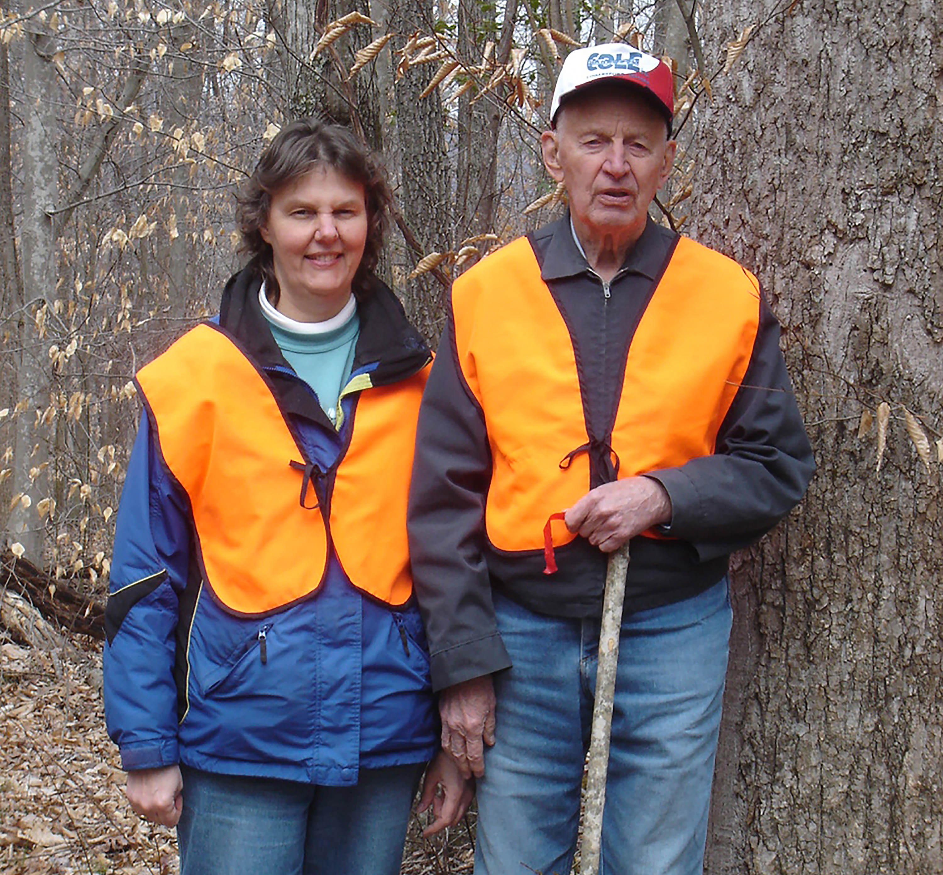 Two people wearing orange vests stand in a forest.