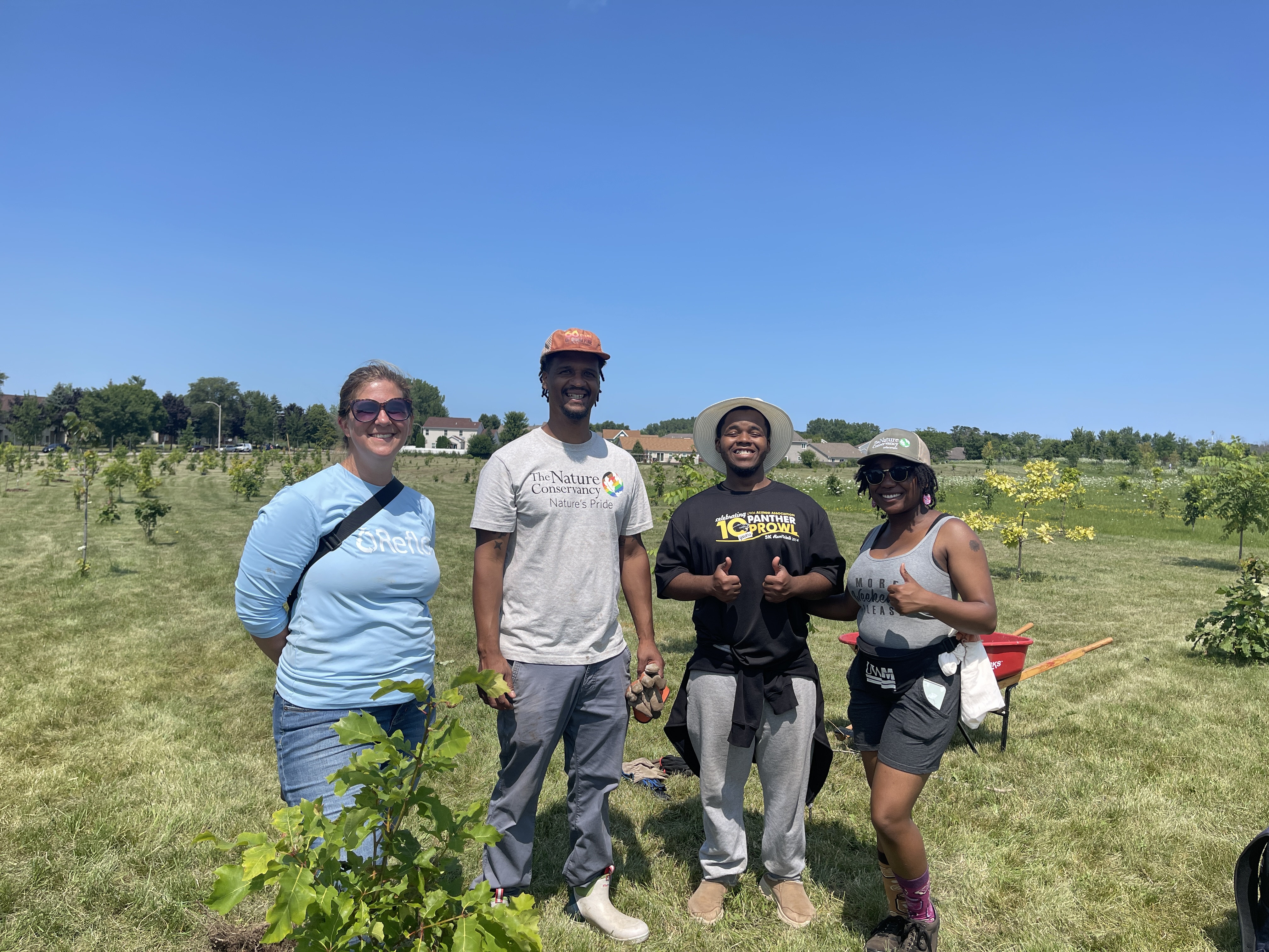 Four people standing outside in a large green space with small, newly planted trees around them.
