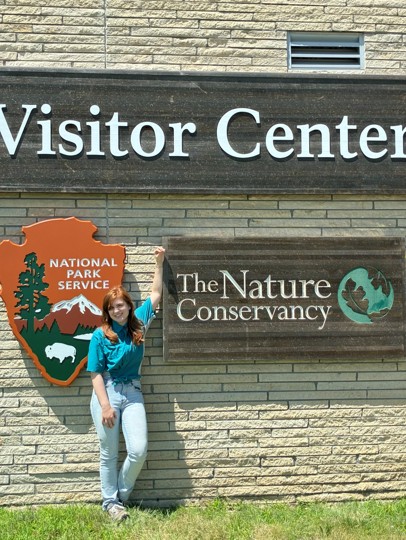 A woman with red hair smiles in front of signage that reads "Visitor Center."