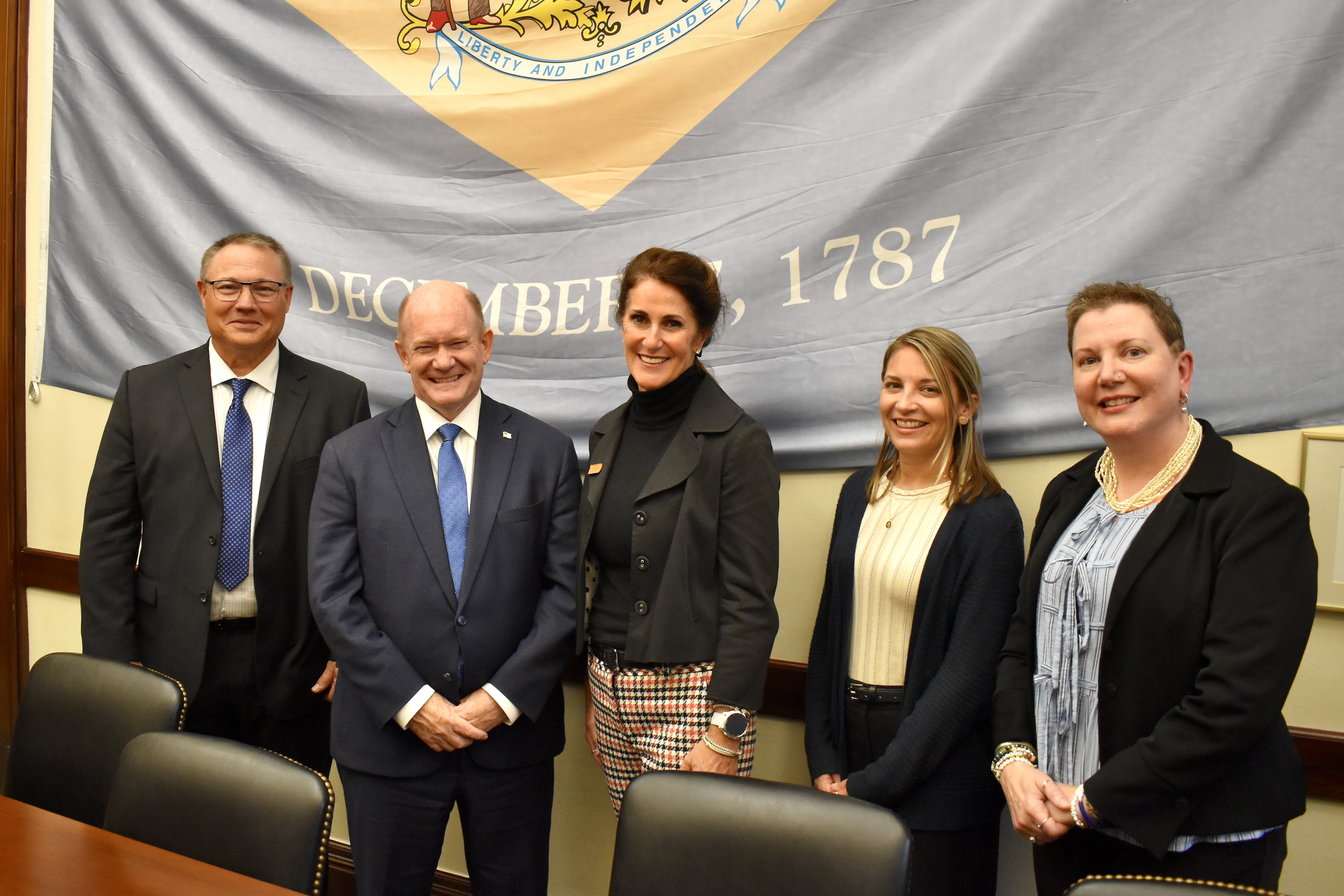 Five people stand in front of a Delaware flag indoors and smile at the camera. 