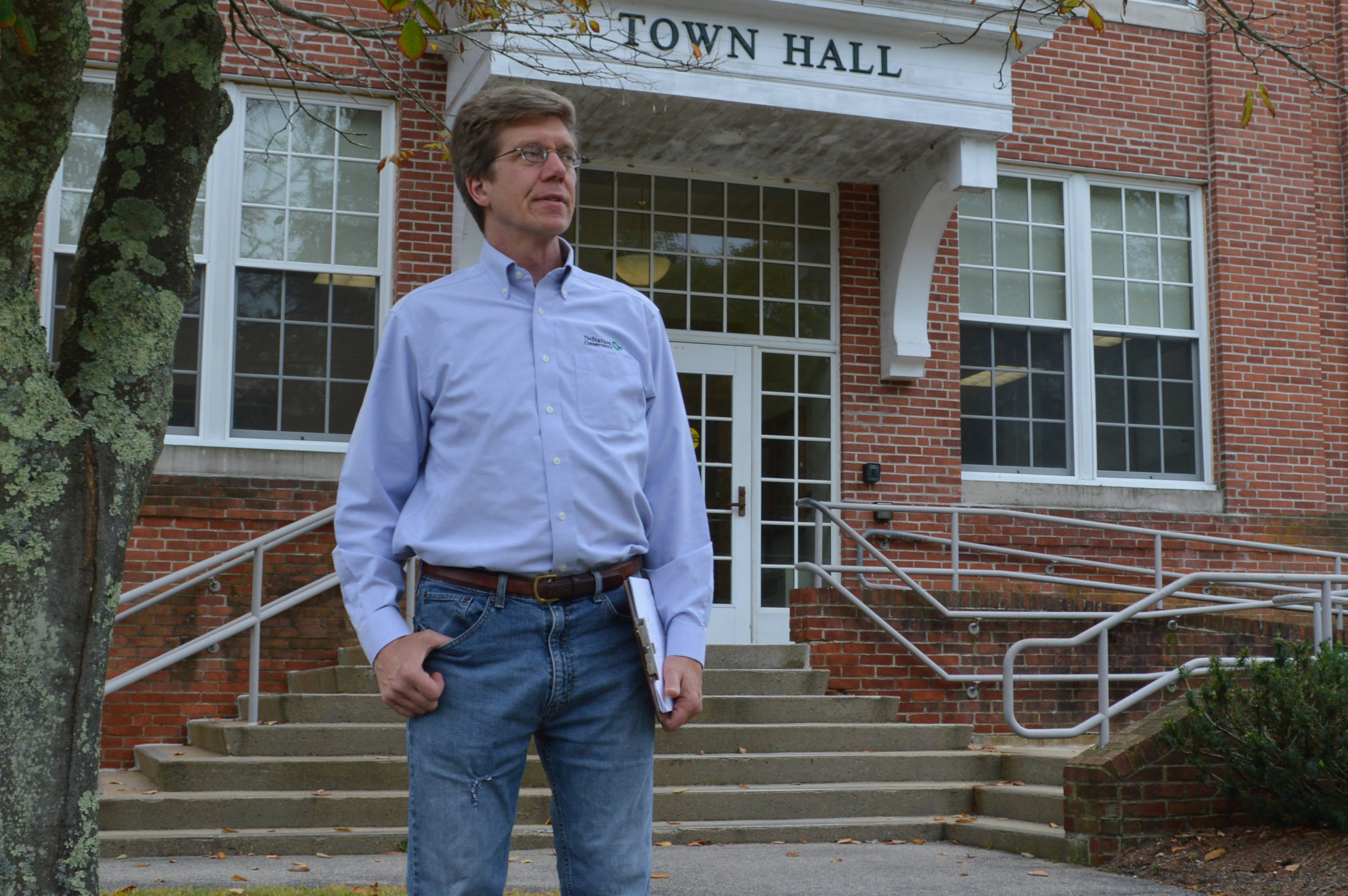 Steve Long stands in front of a town hall building holding a clipboard and looking into the distance.