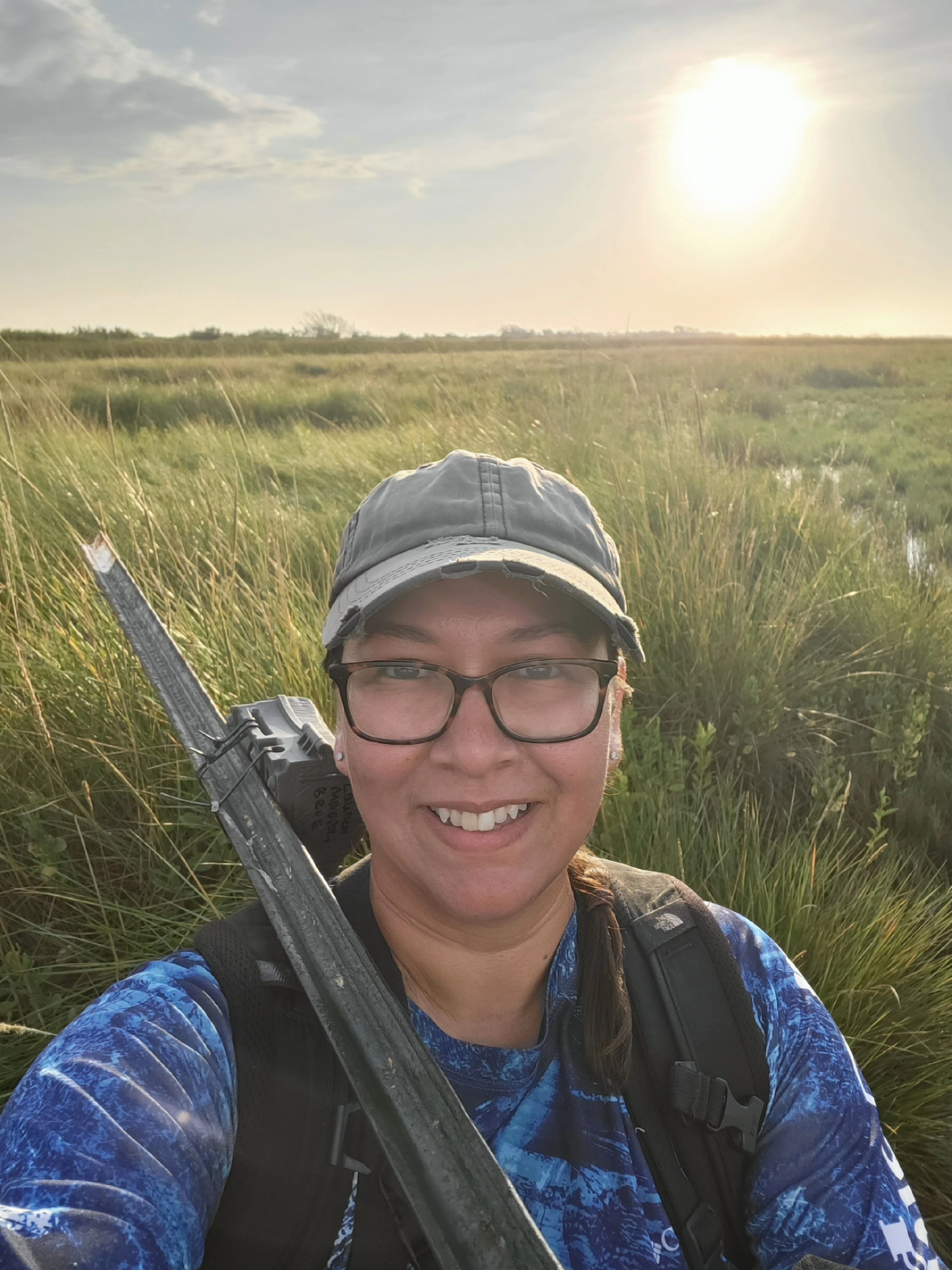 A woman wearing a hat and glasses stands in a field.