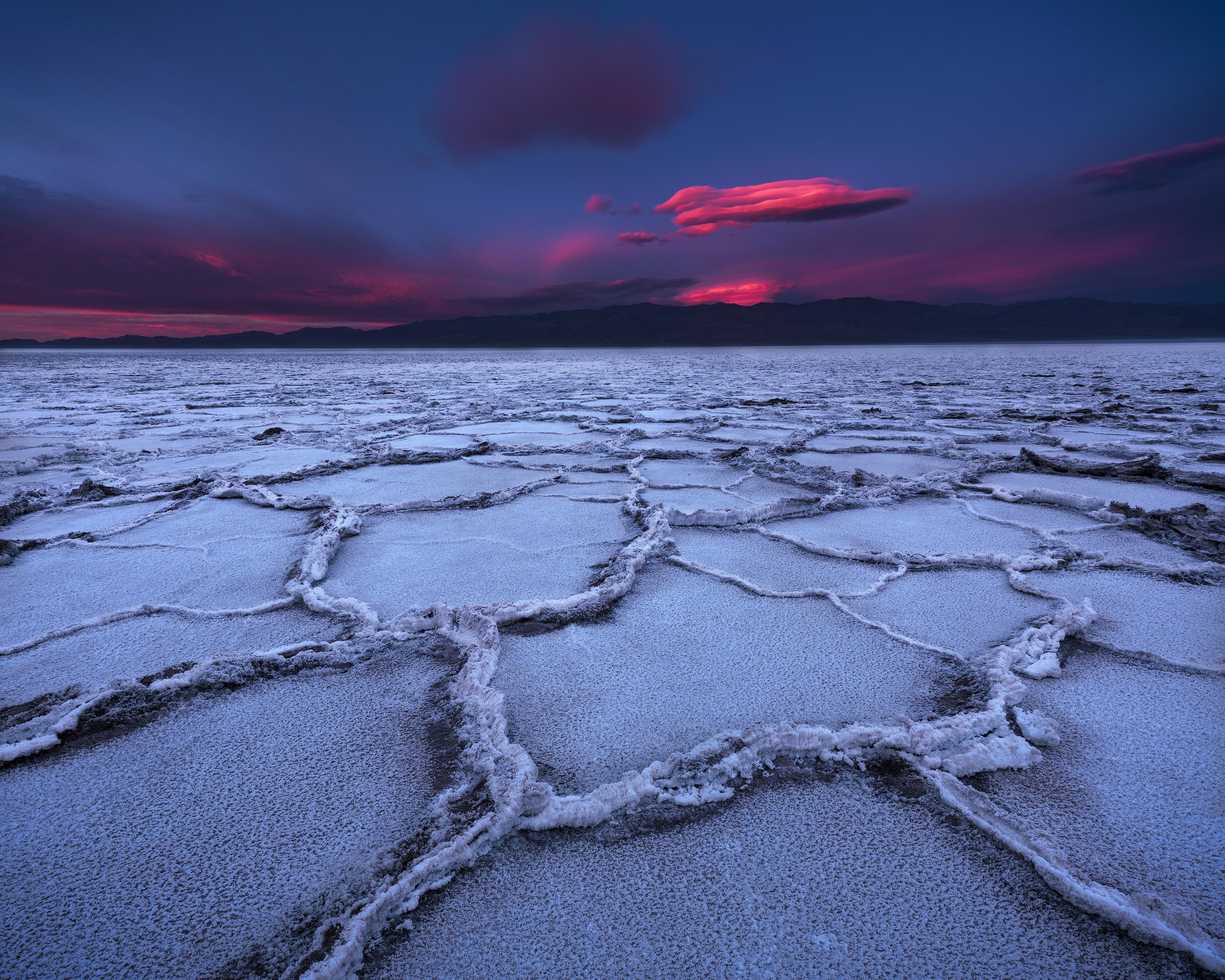 Ground-level view across a mosaic-like salt flat in Death Valley, with a dramatic sky overhead highlighted by vibrant pink clouds.