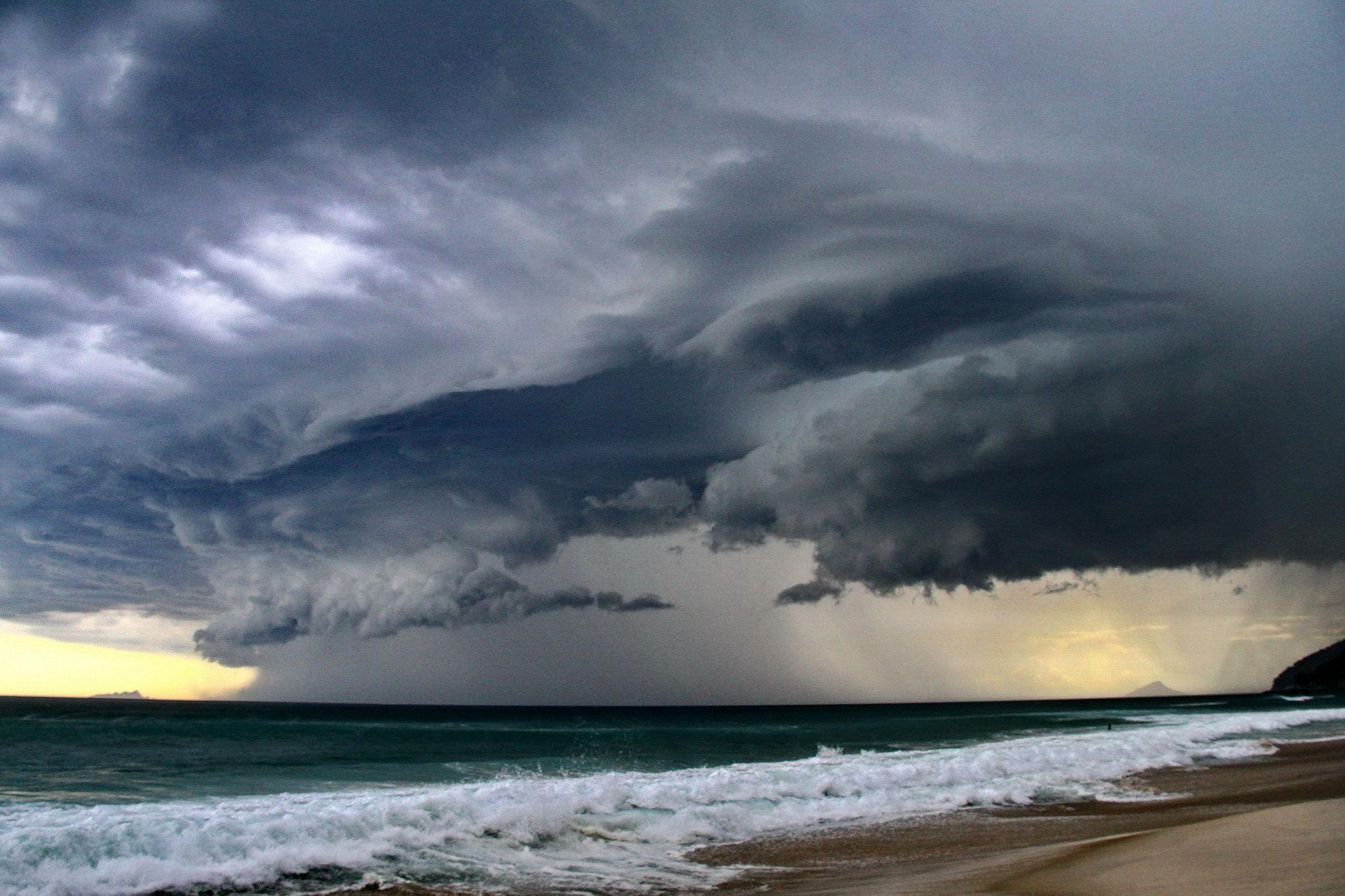 Landscape view of a beach and coastline with dramatic gray storm clouds overhead and sheets of rain pouring down over the ocean in the distance.