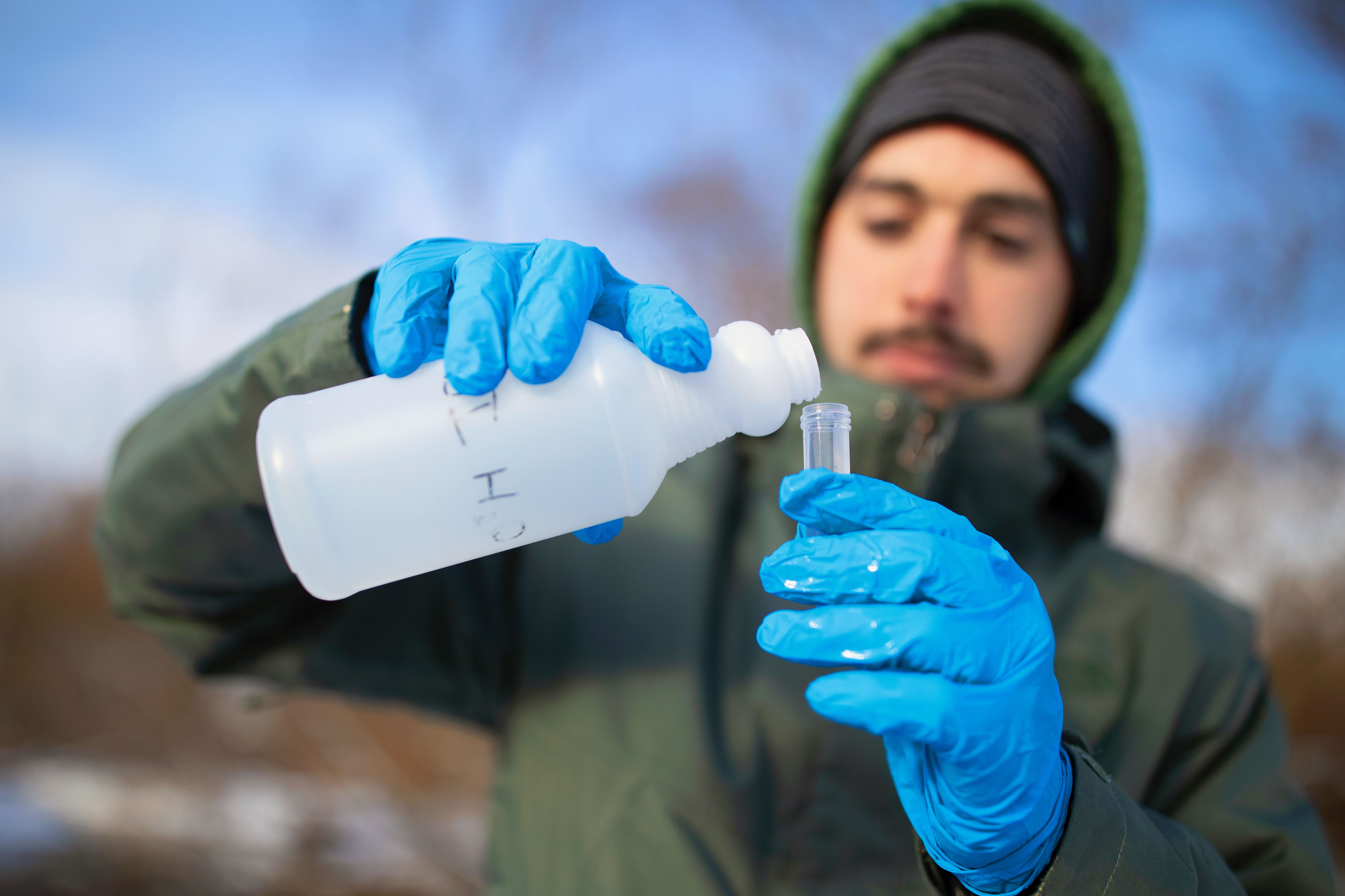 A person dressed in drab green winter outerwear and blue disposable gloves pouring water into a test tube from an opaque plastic bottle.