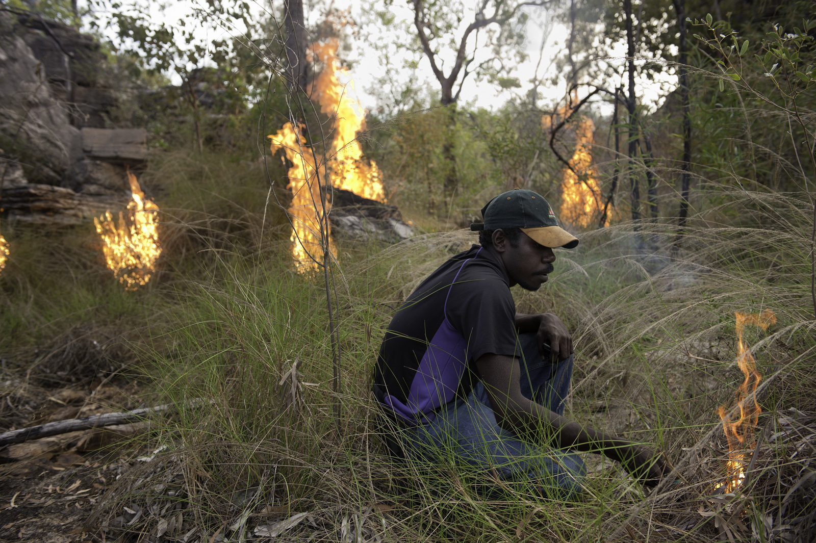 A person sits in grasses among small fires he has set.