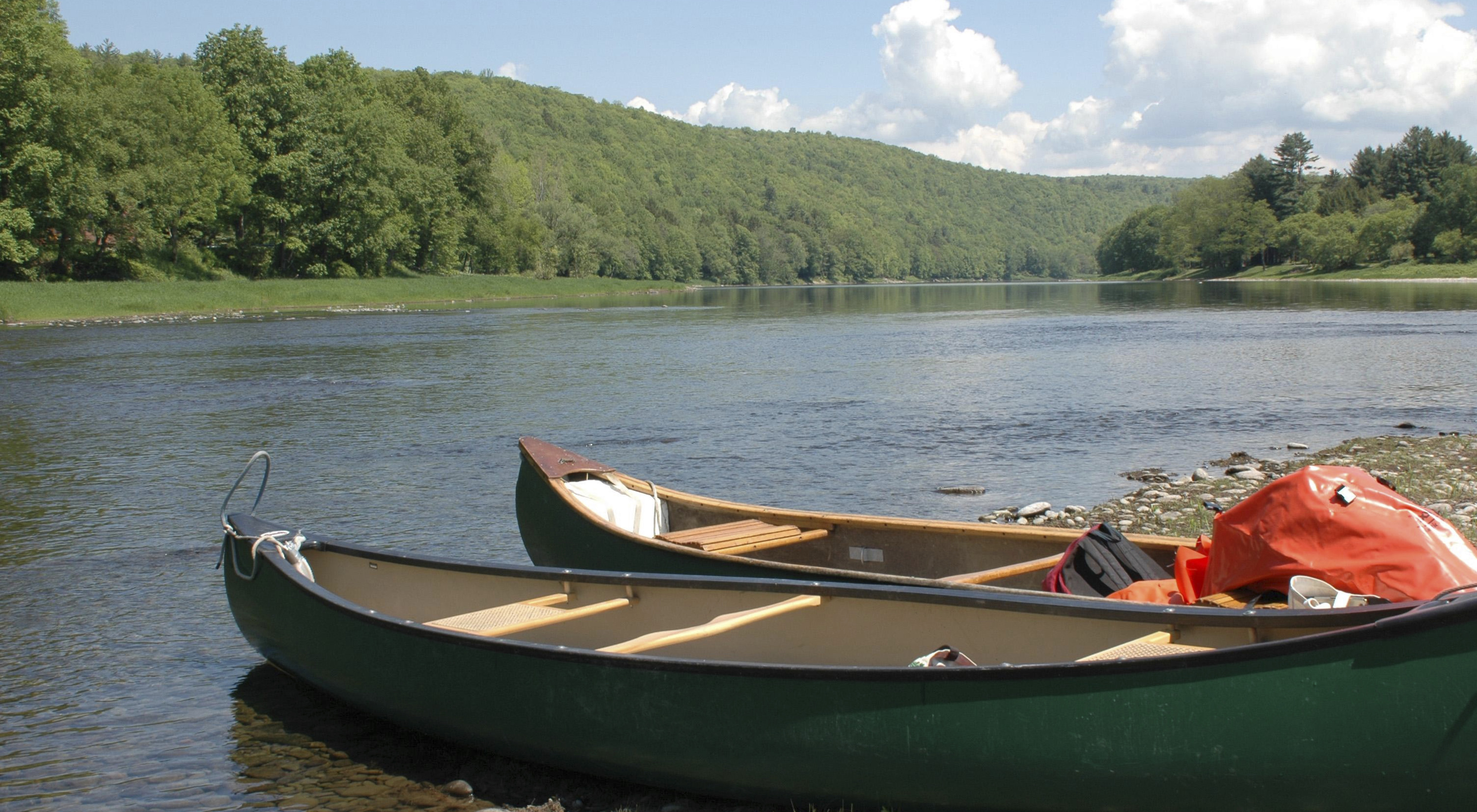 Two canoes sit on the side of a river bank. A tall, tree-covered ridge rises in the background under a blue sky and white puffy clouds.