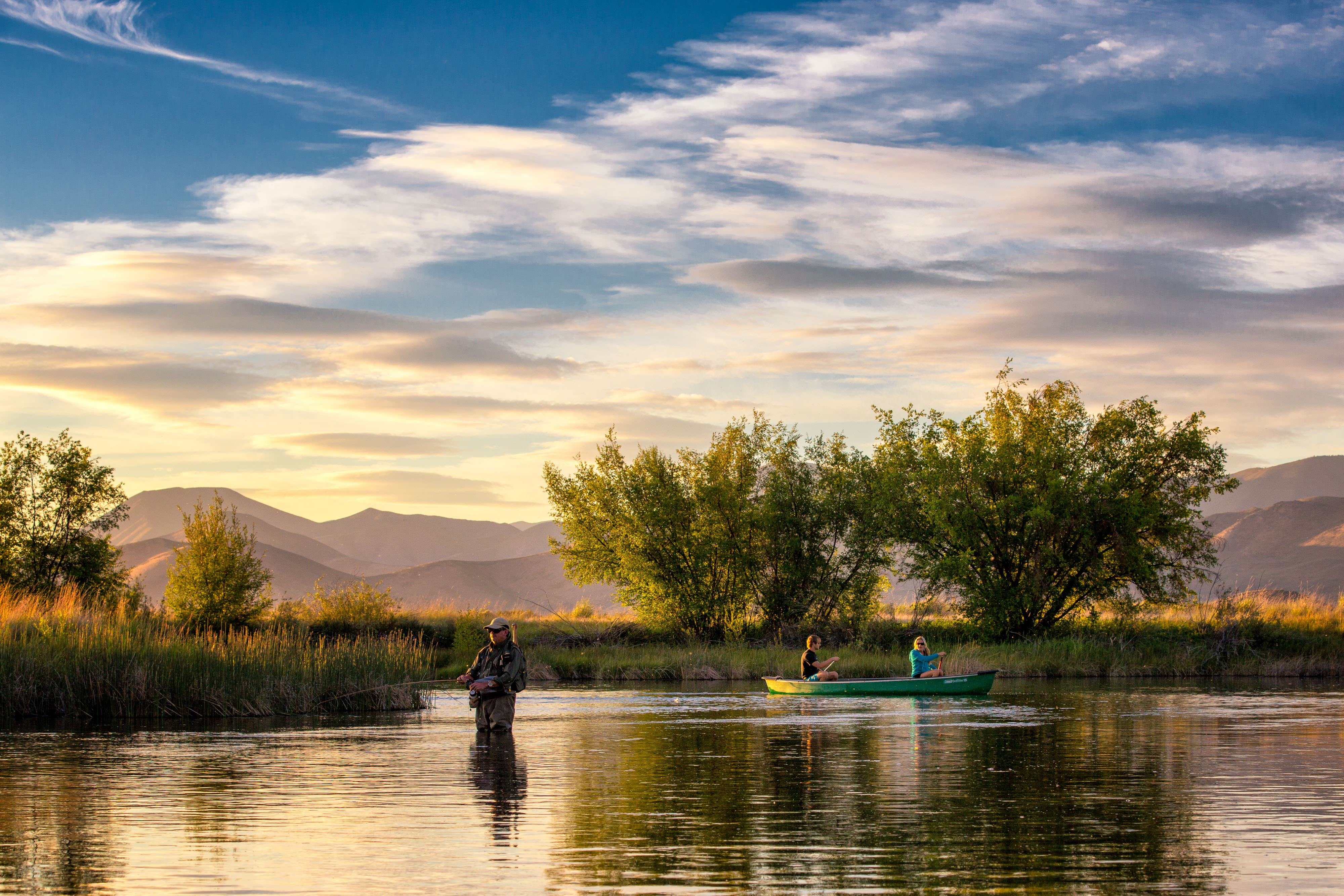 Angler fishes a river with two people in canoe nearby.