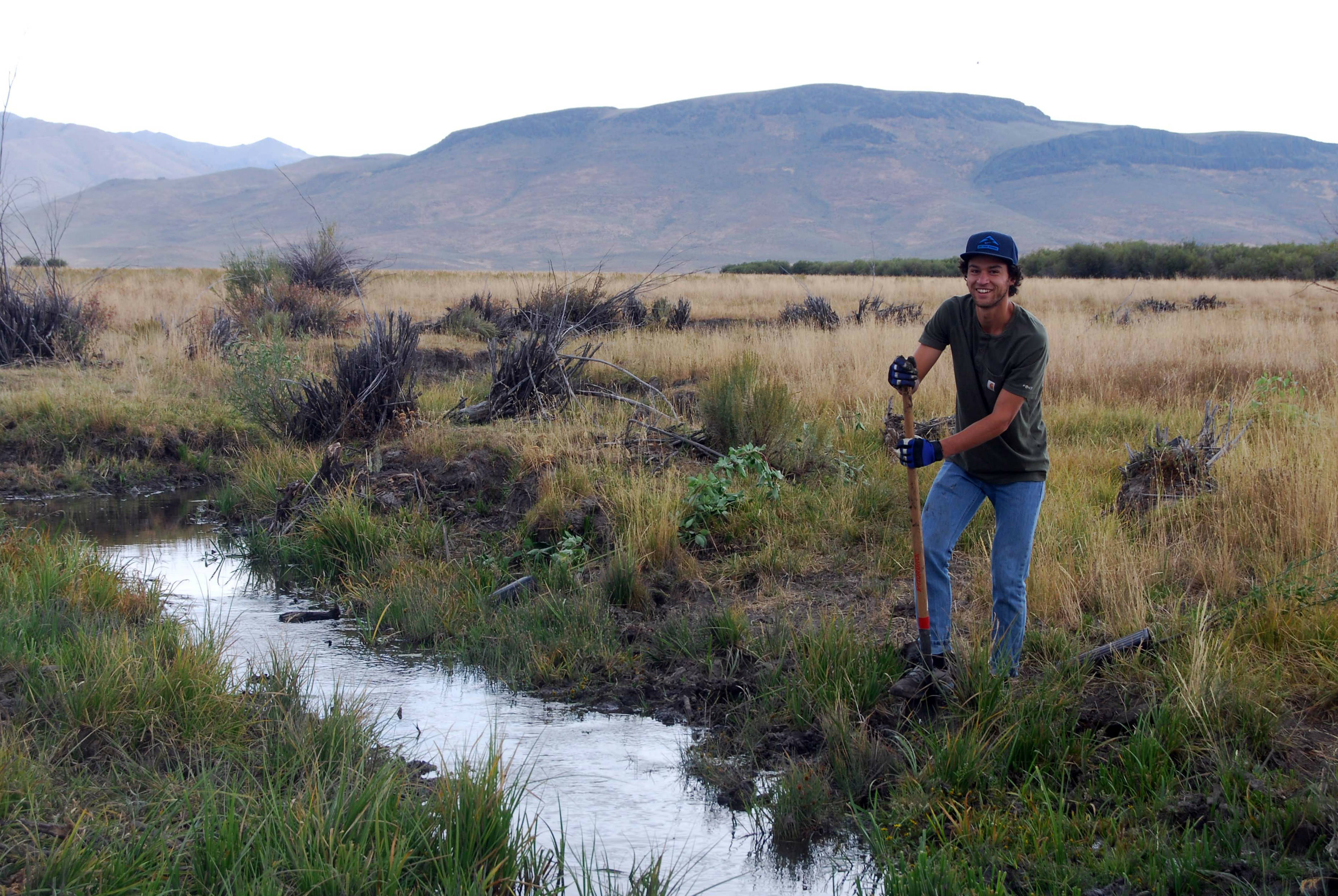 A man with a shovel stands in a large grassy field at the edge of a narrow creek.