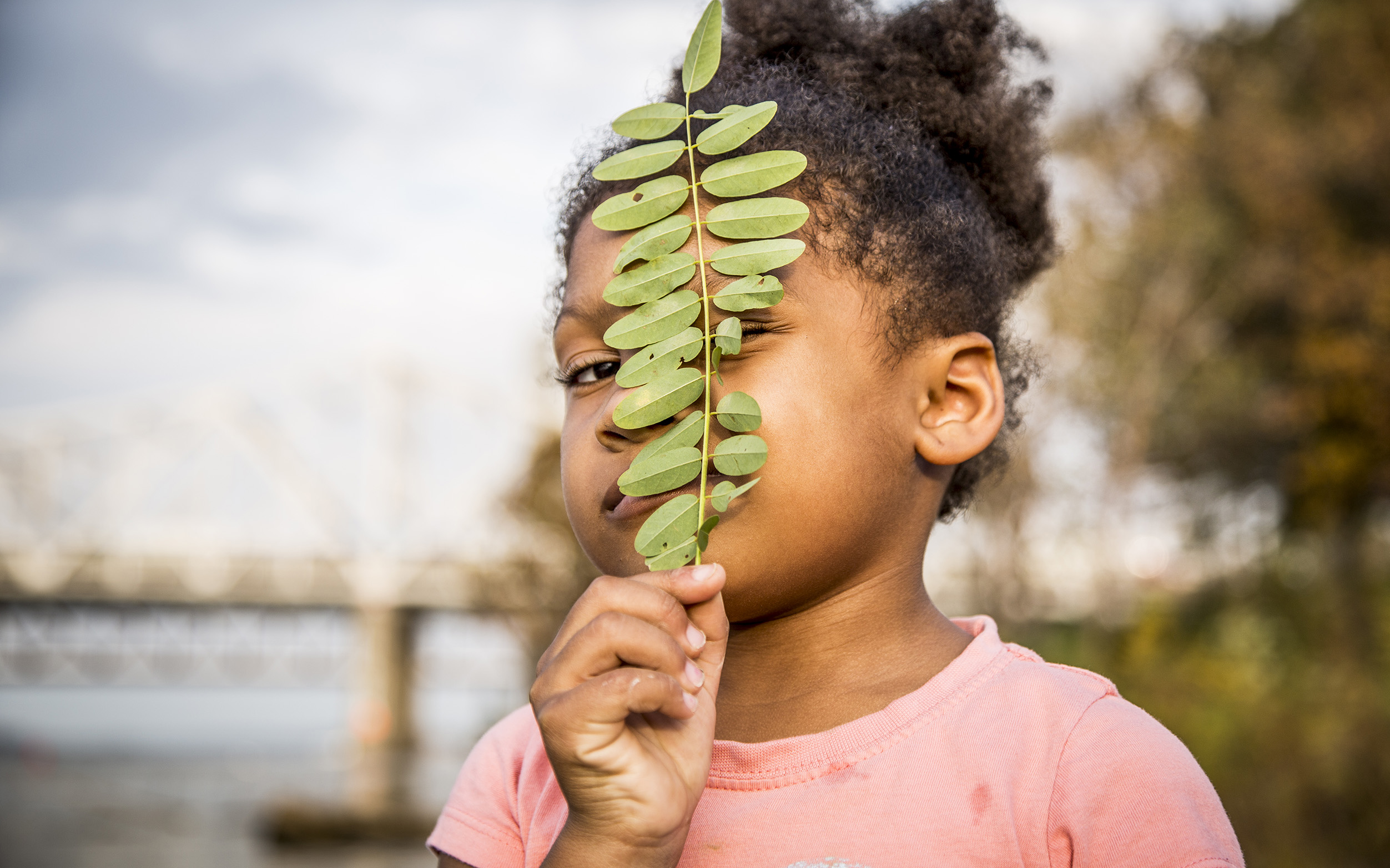A small child in a pink blouse holds a green leaf in front of her face.