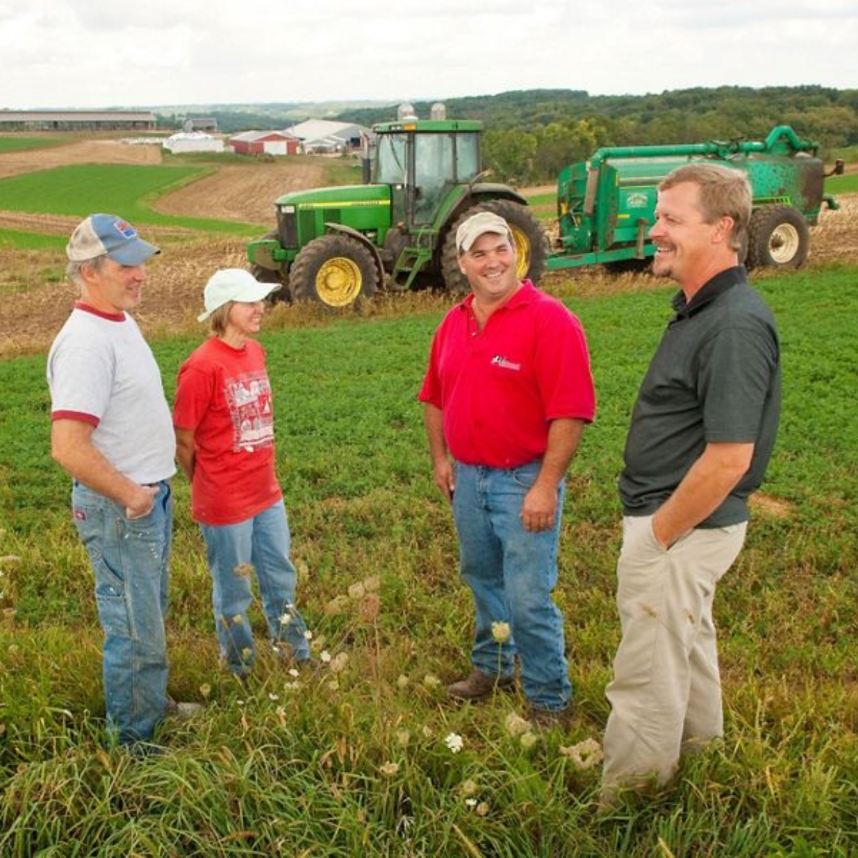 Four people stand in a green agricultural field, gathered in conversation amid tall grass and wildflowers. Behind the group, a large green tractor and attached farm equipment are parked on a gently sloping hillside. The landscape features alternating strips of green crops and brown cultivated soil that curve across rolling terrain. Several farm buildings, including red barns, white agricultural structures, and equipment sheds, are visible in the distance. A tree-covered ridge stretches across the horizon beneath a cloudy, overcast sky. The image highlights a working agricultural landscape and farm management setting.
