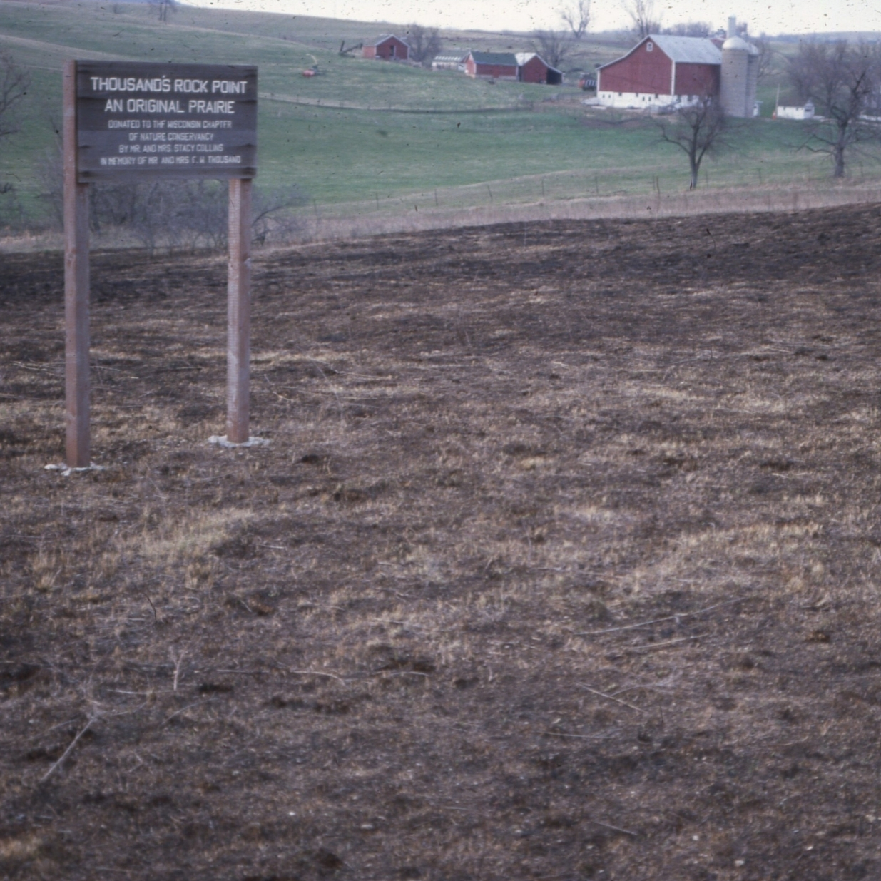 A faded color archival photo of a wooden sign marking Thousand's Rock Point, an Original Prairie stands in a prairie landscape, with rolling farmland, red barns, and a silo visible in the background.